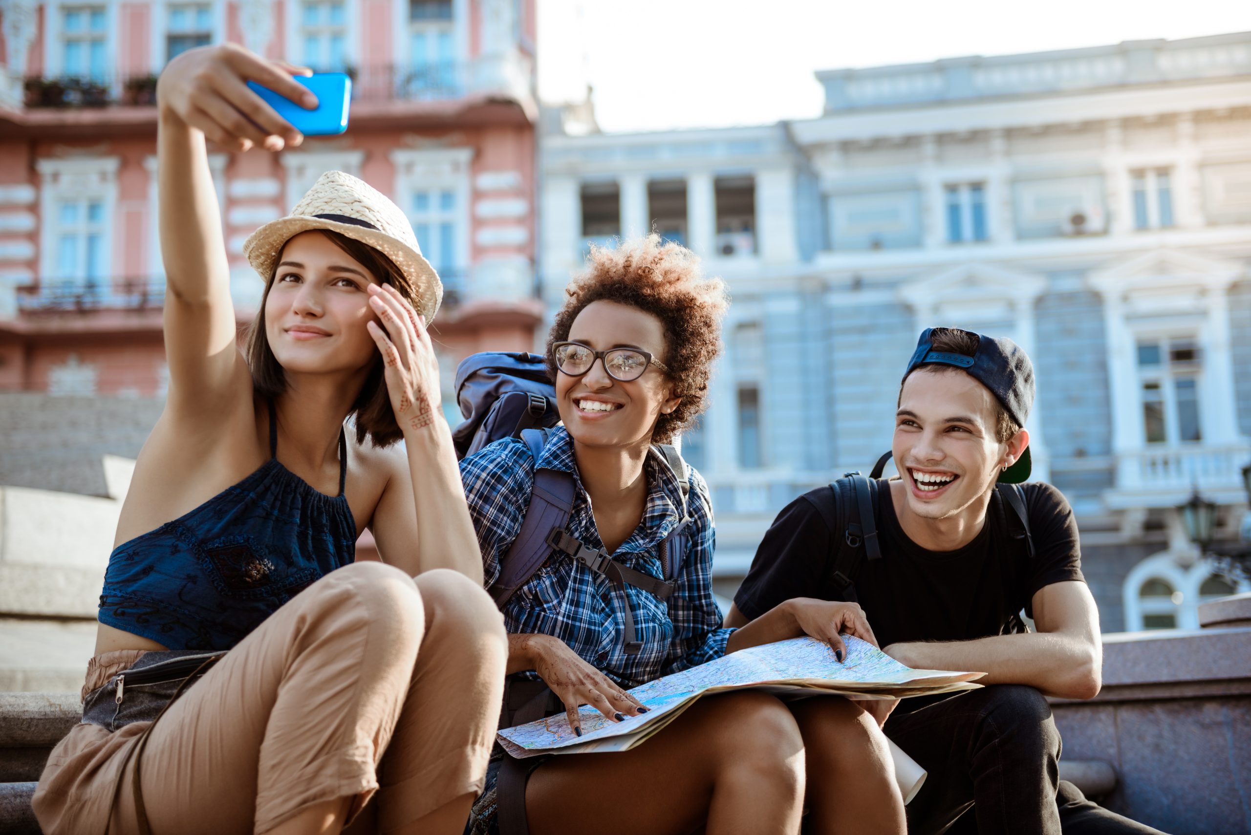 Young beautiful friends travelers with backpacks smiling, making selfie, sitting near sight.