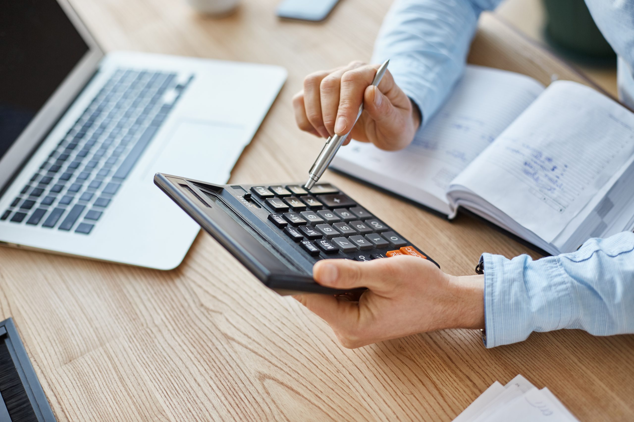 Cropped view of professional serious finance manager, holding calculator in hands, checking company month’s profits, looking through details on laptop, writing down information for meeting with director. Cropped view of professional serious finance manager, holding calculator in hands, checking company month's profits, looking through details on laptop, writing down information for meeting with director