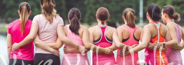 Group of women runners in pink shirts gather in solidarity and prepare to run. They are facing away from the camera toward a pond and are embracing each other.
