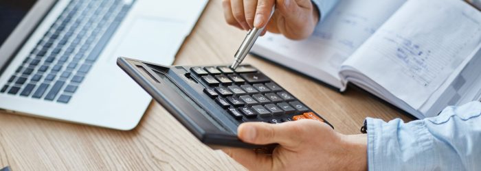 Cropped view of professional serious finance manager, holding calculator in hands, checking company month's profits, looking through details on laptop, writing down information for meeting with director