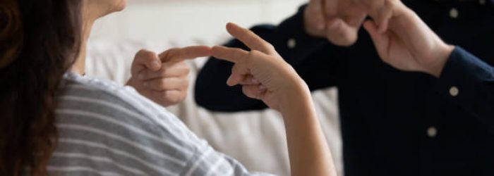 Young positive couple sit on sofa at home communicating using sign language, friends talking indoor showing fingers gestures, having fun, leading pleasant conversation, people with disability concept