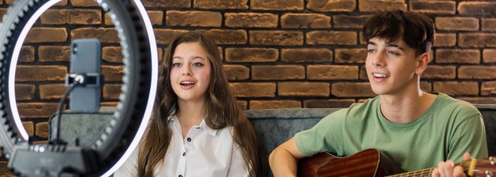 Young smiling man and woman playing on a guitar and singing, shooting themselves on smartphone on a tripod with ring light in a studio